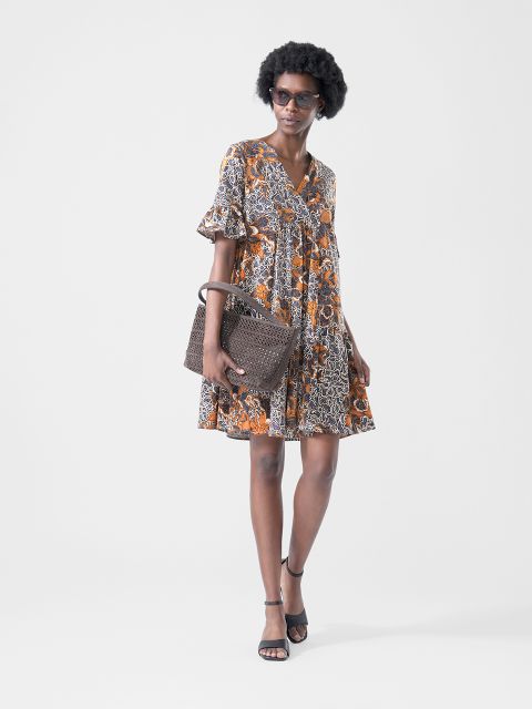 Une femme porte une robe courte à motifs dans les tons orange et noir, des lunettes de soleil, des sandales noires et tient un sac à main marron texturé.