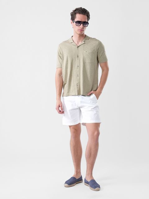 Un homme portant des lunettes de soleil, une chemise à manches courtes vert olive clair, un short blanc et des espadrilles bleu marine se tient devant un simple fond blanc.