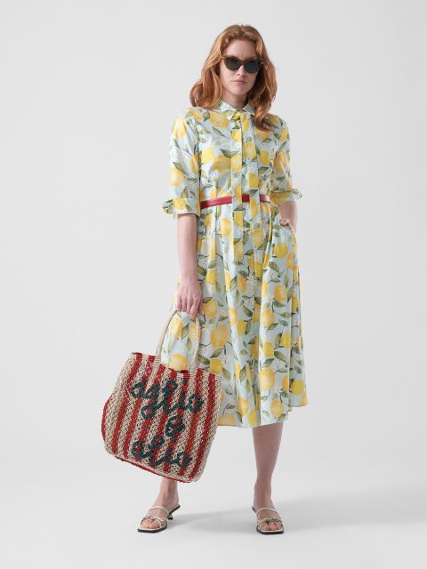 Femme vêtue d'une robe à motif de citrons, portant des lunettes de soleil et des sandales, tenant un sac rayé rouge et blanc avec une inscription, devant un fond blanc uni.