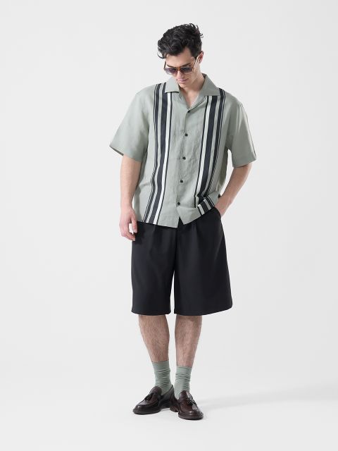 Un homme portant des lunettes de soleil, une chemise vert clair à manches courtes avec des rayures longitudinales noires, un short noir, des chaussettes vertes et des chaussures basses marron, regarde vers le bas.
