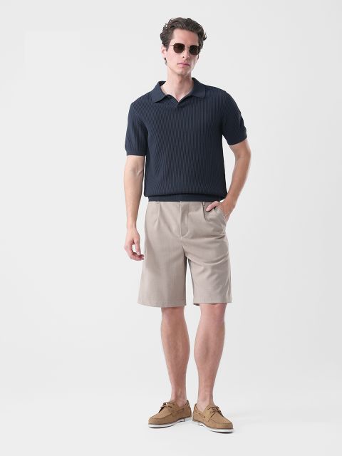 Homme avec des lunettes de soleil, un polo bleu marine à manches courtes, un short beige arrivant aux genoux et des chaussures bateau marron, devant un simple fond blanc.