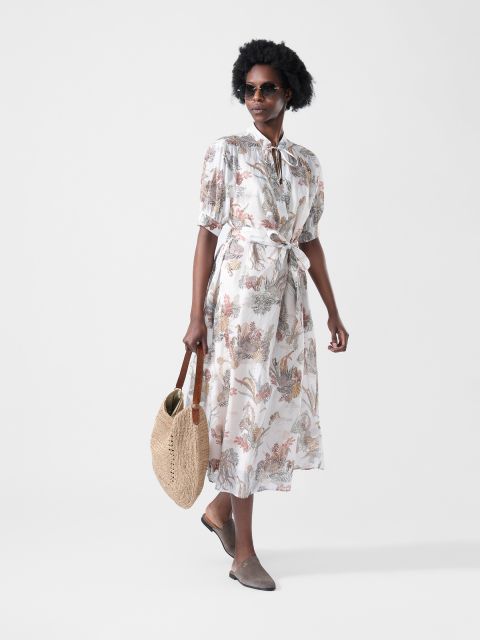 Femme en robe midi fleurie avec un lien à la taille, des lunettes de soleil, un sac tissé à la main et des mocassins marron.
