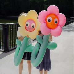 Deux personnes tiennent de grandes fleurs gonflables aux visages souriants, masquant leurs propres visages, alors qu'elles se tiennent sur un pont au bord de l'eau.
