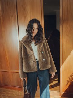 Une femme aux cheveux bouclés, portant un manteau en fausse fourrure marron, un chemisier blanc et un jean à jambes larges, se tient à l'intérieur avec un sac marron à la main, avec des panneaux de bois et la lumière du soleil en arrière-plan.
