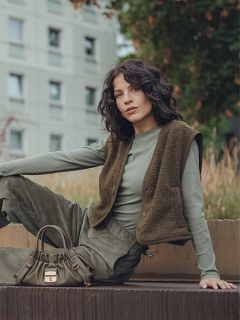 Une femme aux cheveux ondulés et foncés est assise à l'extérieur sur une surface surélevée, portant un haut côtelé vert à manches longues, un gilet olive et un pantalon assorti. Elle a un petit sac à main olive à côté d'elle et un immeuble résidentiel en arrière-plan.