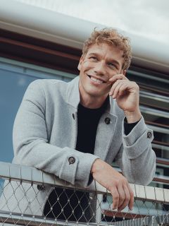 Un homme souriant aux cheveux blonds bouclés est nonchalamment appuyé contre une clôture en grillage, portant un manteau gris clair sur une chemise noire, avec un bâtiment moderne en arrière-plan.