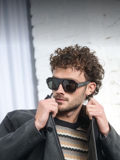 Un homme avec des lunettes de soleil, une veste en cuir noire et un pull rayé se tient devant un mur de briques blanches et ajuste le col de sa veste d'une main.