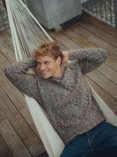 Un jeune homme aux cheveux blonds bouclés, vêtu d'un pull marron et d'un jean, se détend dans un hamac, sourit et regarde sur le côté. Le hamac est installé sur une terrasse en bois avec une balustrade métallique en arrière-plan.