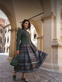 Une femme portant une jupe à motifs et une veste verte se tient sous des arches de pierre, souriant et tenant un sac à main vert.