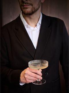 Un homme en costume noir et chemise blanche tient un verre à champagne transparent et nervuré à la main, son visage étant partiellement hors du cadre.