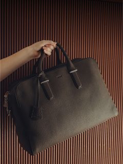 Une main tient une grande serviette en cuir noir avec deux poignées devant un fond brun rainuré.