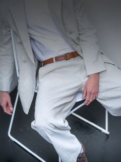 Une personne en costume gris clair et chemise blanche est assise sur une chaise blanche, une ceinture marron est visible et son visage n'est pas visible.