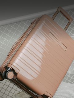 Une valise rigide brillante, brun clair, avec des roulettes et une poignée extensible est posée sur un sol carrelé.