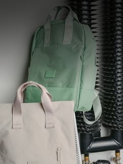Un sac à dos vert menthe et un sac fourre-tout beige sont suspendus devant des tuyaux et des bobines noirs sur un mur.