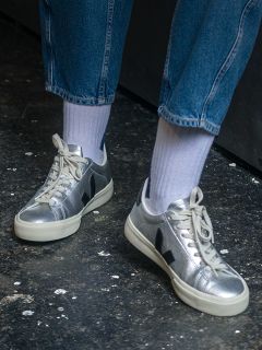 Personne portant des baskets métallisées argentées avec des semelles blanches, des chaussettes blanches et un jean bleu, debout sur un sol sombre et texturé.