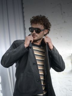 Homme aux cheveux bouclés portant des lunettes de soleil, un pull rayé et une veste en cuir noire, ajustant son col devant un mur de briques blanches.