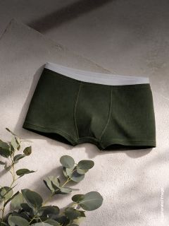 Des boxershorts vert foncé pour hommes avec une ceinture blanche sont exposés sur une surface texturée, avec des feuilles d'eucalyptus à proximité dans une lumière douce et naturelle.