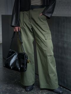 Personne portant un pantalon cargo vert olive, des chaussures noires et une veste noire, tenant un sac à main noir brillant à ses côtés.