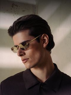 Un jeune homme aux cheveux foncés, portant des lunettes de soleil marron et une chemise foncée à col, regarde sur le côté dans une lumière douce et ombragée.