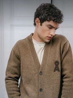 Un homme porte un cardigan marron boutonné avec un emblème brodé foncé sur la poitrine par-dessus un t-shirt crème.