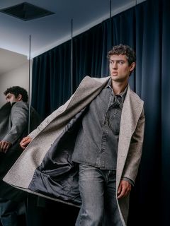 Un homme portant une chemise grise à col et un jean enfile un long manteau de laine de couleur neutre.